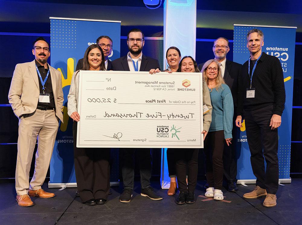 Five college-age startup founders pose onstage with two sponsors, two hosts, and a giant check for $25,000