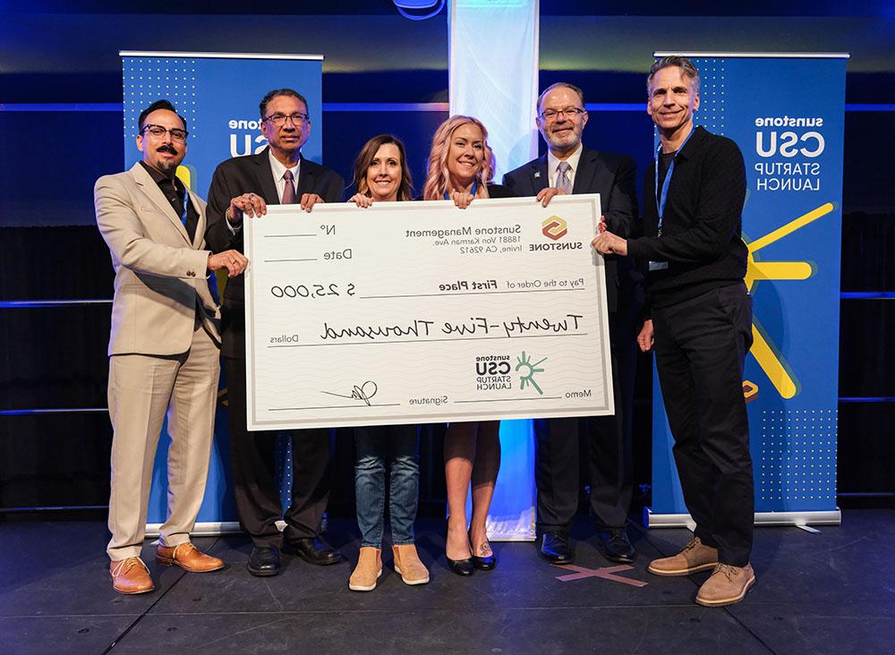 Two female, college-age startup founders pose onstage with two sponsors, two hosts, and a giant check for $25,000