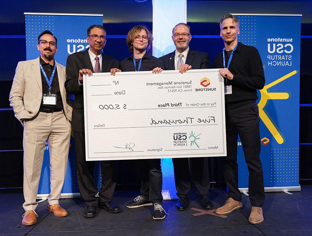 A male, college-age startup founder poses onstage with two sponsors, two hosts, and a giant check for $5,000