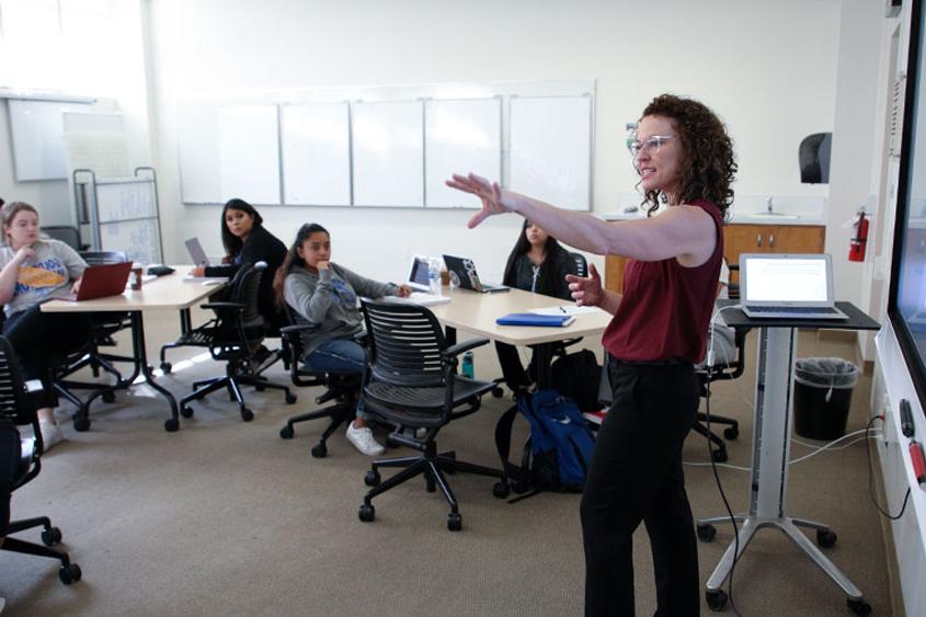 A professor with their arm extended out while talking to the classroom.